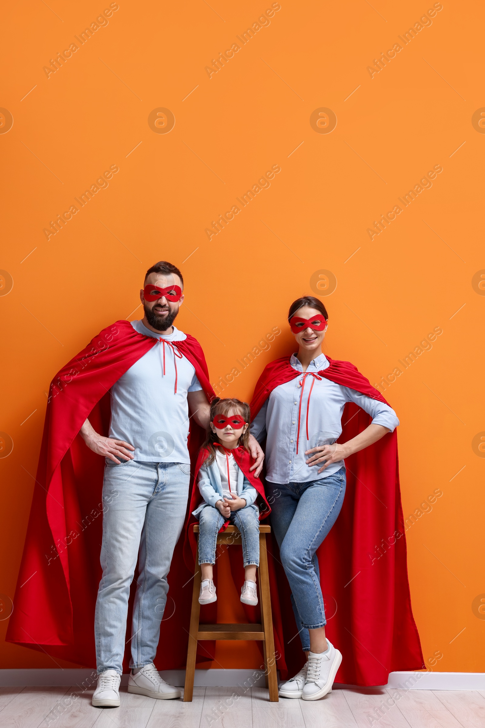 Photo of Parents and their cute little daughter wearing superhero costumes near orange wall indoors, space for text