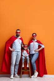 Parents and their cute little daughter wearing superhero costumes near orange wall indoors, space for text