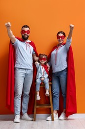 Photo of Parents and their cute little daughter wearing superhero costumes near orange wall indoors