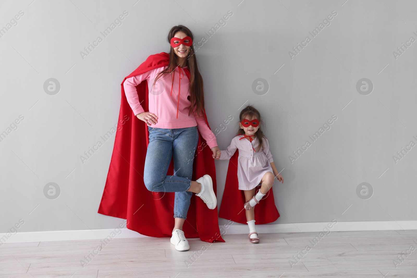 Photo of Mother and her cute little daughter wearing superhero costumes near gray wall indoors