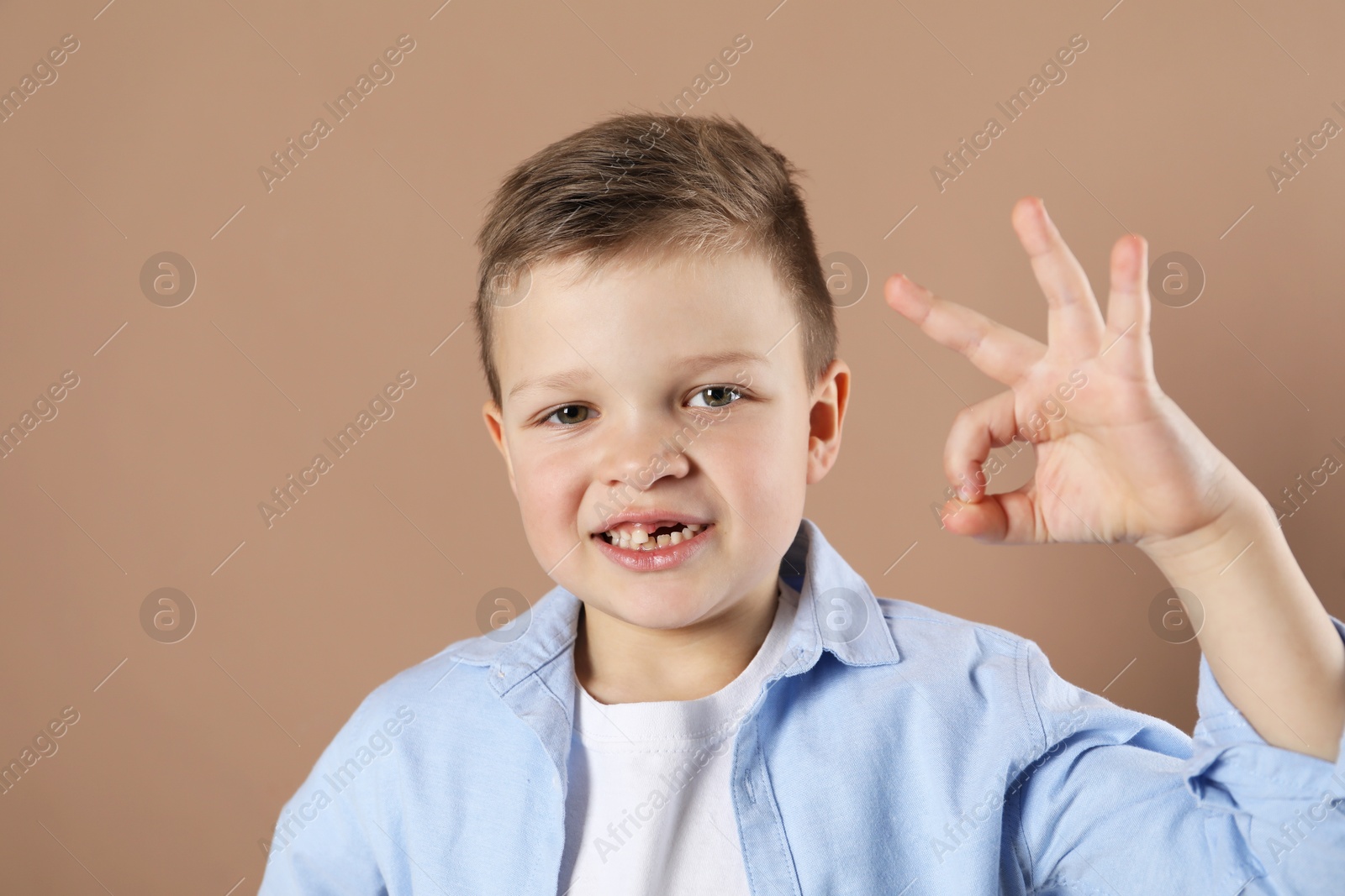 Photo of Cute little boy with missing tooth showing ok gesture on dark beige background