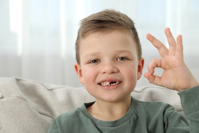 Photo of Cute little boy with missing tooth showing ok gesture at home