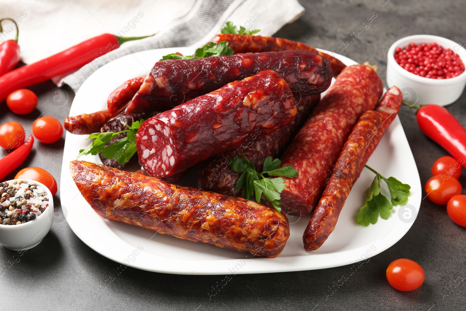 Photo of Different smoked sausages, parsley, peppercorns, tomatoes and chili pepper on grey textured table, closeup