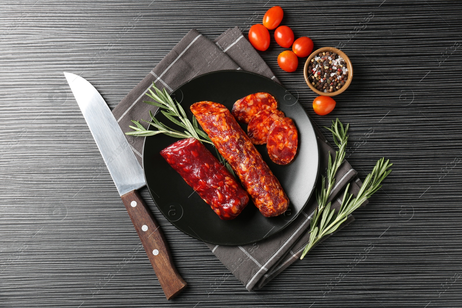 Photo of Cut smoked sausages, rosemary, peppercorns and tomatoes on black wooden table, flat lay