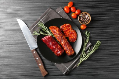 Photo of Cut smoked sausages, rosemary, peppercorns and tomatoes on black wooden table, flat lay