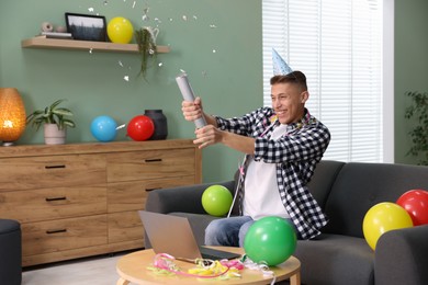 Happy man blowing up confetti popper on sofa in room decorated for party