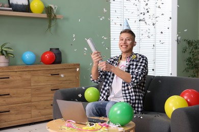 Photo of Happy man blowing up confetti popper on sofa in room decorated for party