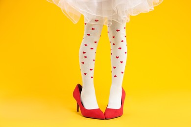 Photo of Little girl wearing oversized high heeled shoes on orange background, closeup