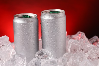 Photo of Refreshing drinks in cans on ice cubes against dark red background, closeup