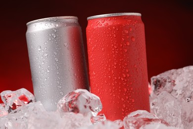 Photo of Refreshing drinks in cans on ice cubes against dark red background, closeup