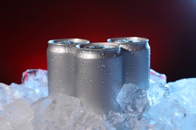 Photo of Refreshing drinks in cans on ice cubes against dark red background, closeup