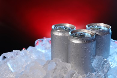 Photo of Refreshing drinks in cans on ice cubes against dark red background, closeup