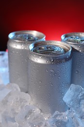 Photo of Refreshing drinks in cans on ice cubes against dark red background, closeup