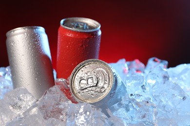 Photo of Refreshing drinks in cans on ice cubes against dark red background, closeup