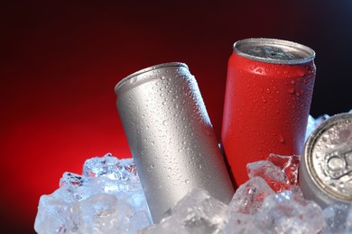 Photo of Refreshing drinks in cans on ice cubes against dark red background, closeup