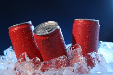 Photo of Refreshing drinks in cans on ice cubes against dark blue background, closeup