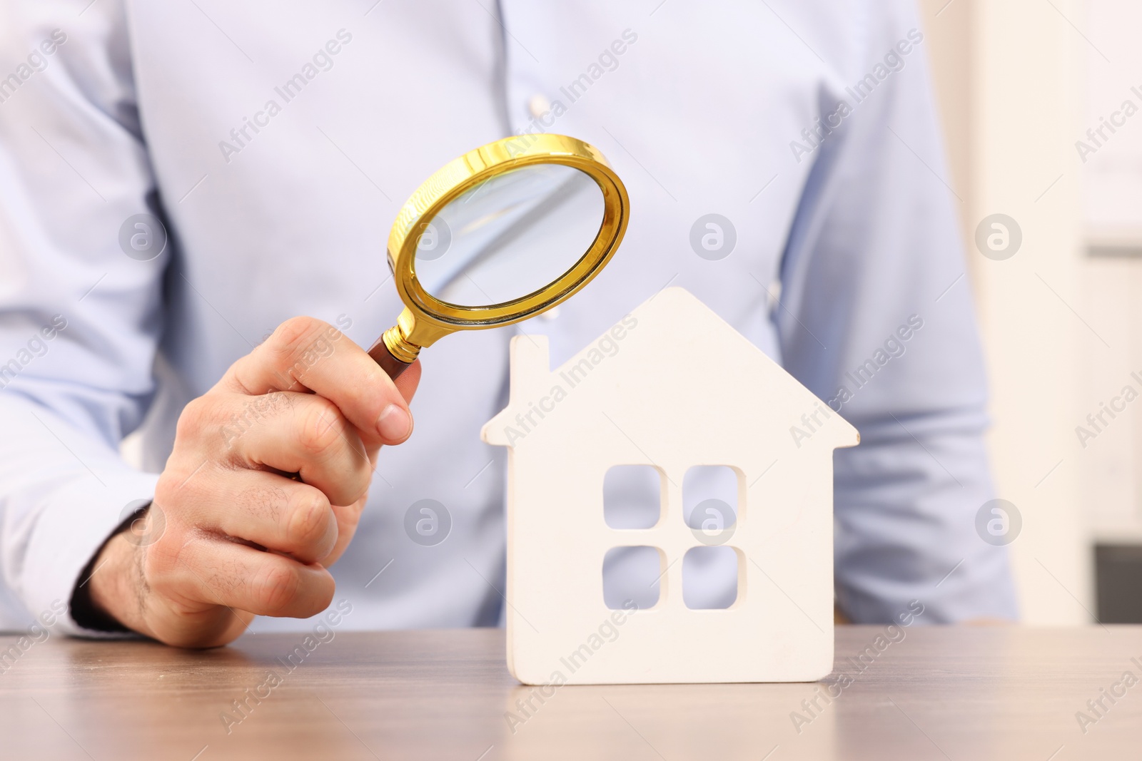 Photo of Real estate agent looking at house figure through magnifying glass at wooden table in office, closeup. Home appraisal