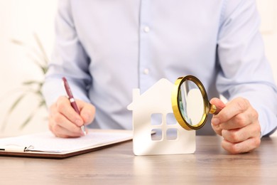 Photo of Real estate agent with house figure and magnifying glass signing document at wooden table in office, closeup. Home appraisal