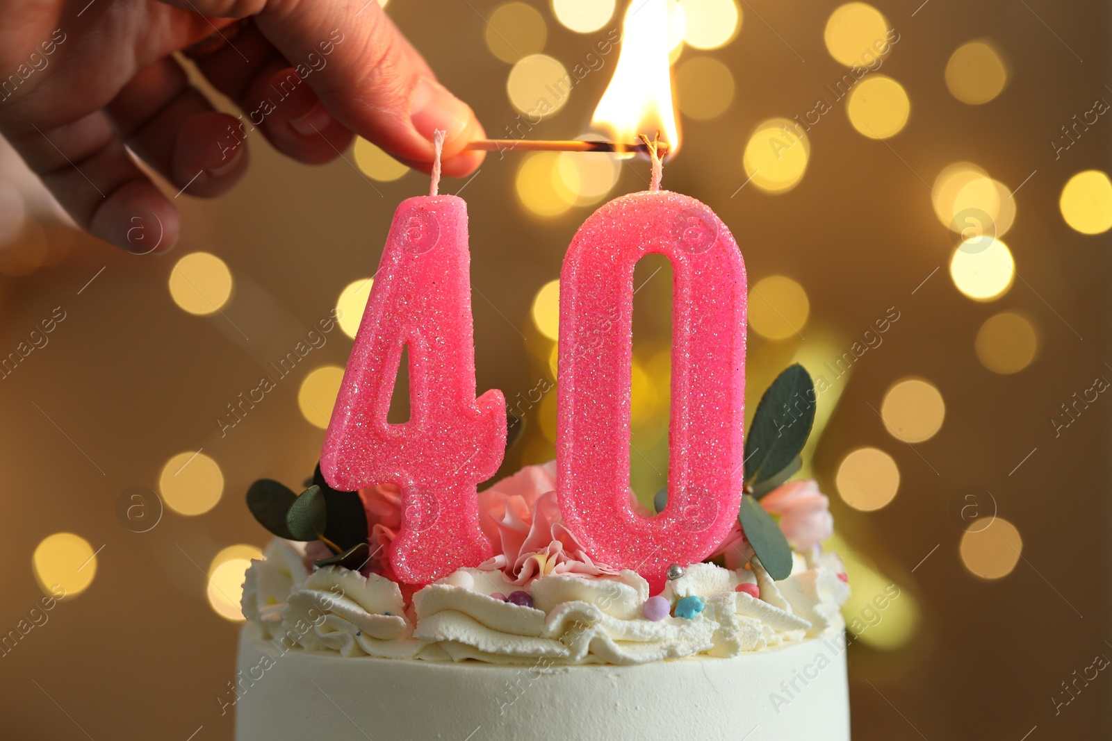 Photo of 40th birthday. Woman lighting number shaped candles on cake against blurred background with lights, closeup. Bokeh effect