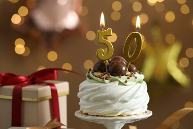 Photo of 50th birthday. Tasty cupcake with burning number shaped candles and gift box on blurred background with lights, closeup. Bokeh effect