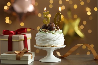 Photo of 50th birthday. Tasty cupcake with burning number shaped candles and gift boxes on wooden table against blurred background with lights, bokeh effect