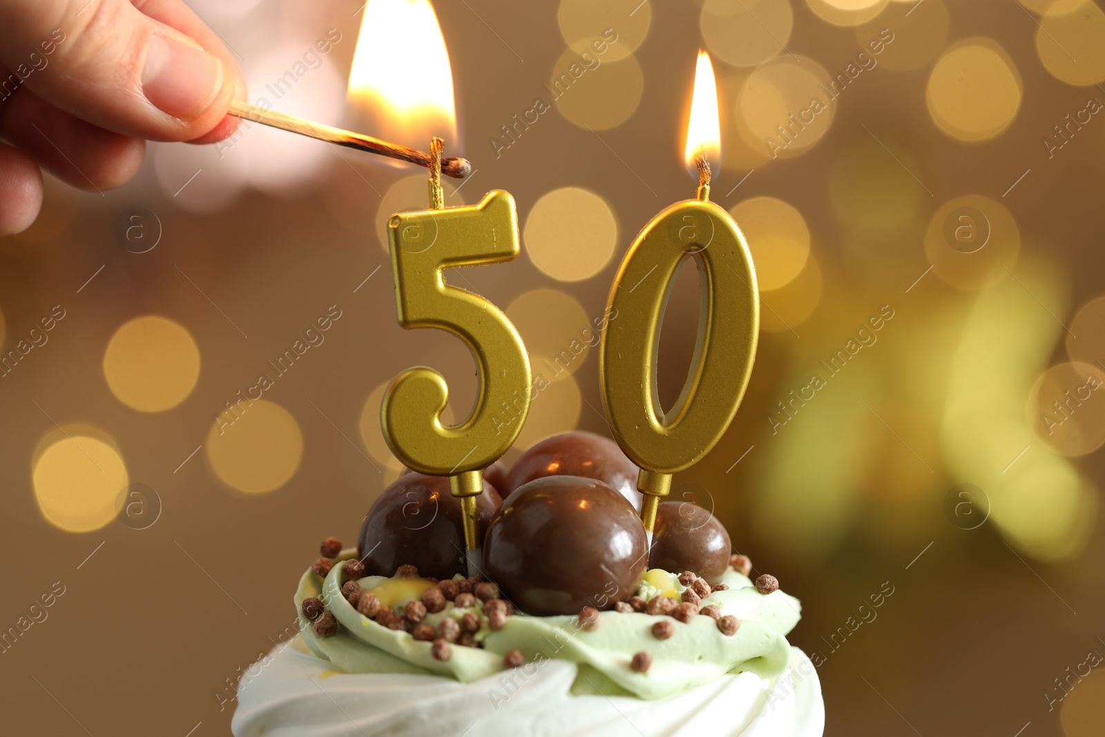 Photo of 50th birthday. Woman lighting number shaped candles on cupcake against blurred background with lights, closeup. Bokeh effect