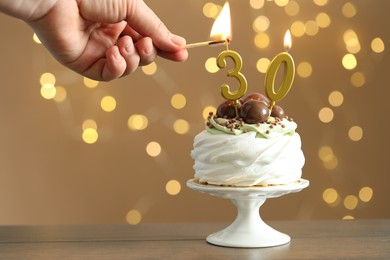 Photo of 30th birthday. Woman lighting number shaped candles on cupcake at wooden against blurred background with lights, closeup. Bokeh effect