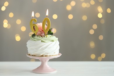 Photo of 60th birthday. Tasty cupcake with burning number shaped candles on white wooden table against grey background with blurred lights, space for text. Bokeh effect