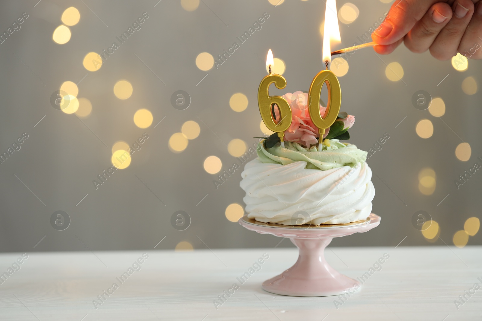 Photo of 60th birthday. Woman lighting number shaped candles on cupcake at white wooden against grey background with blurred lights, closeup. Space for text