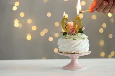 Photo of 60th birthday. Woman lighting number shaped candles on cupcake at white wooden against grey background with blurred lights, closeup. Space for text