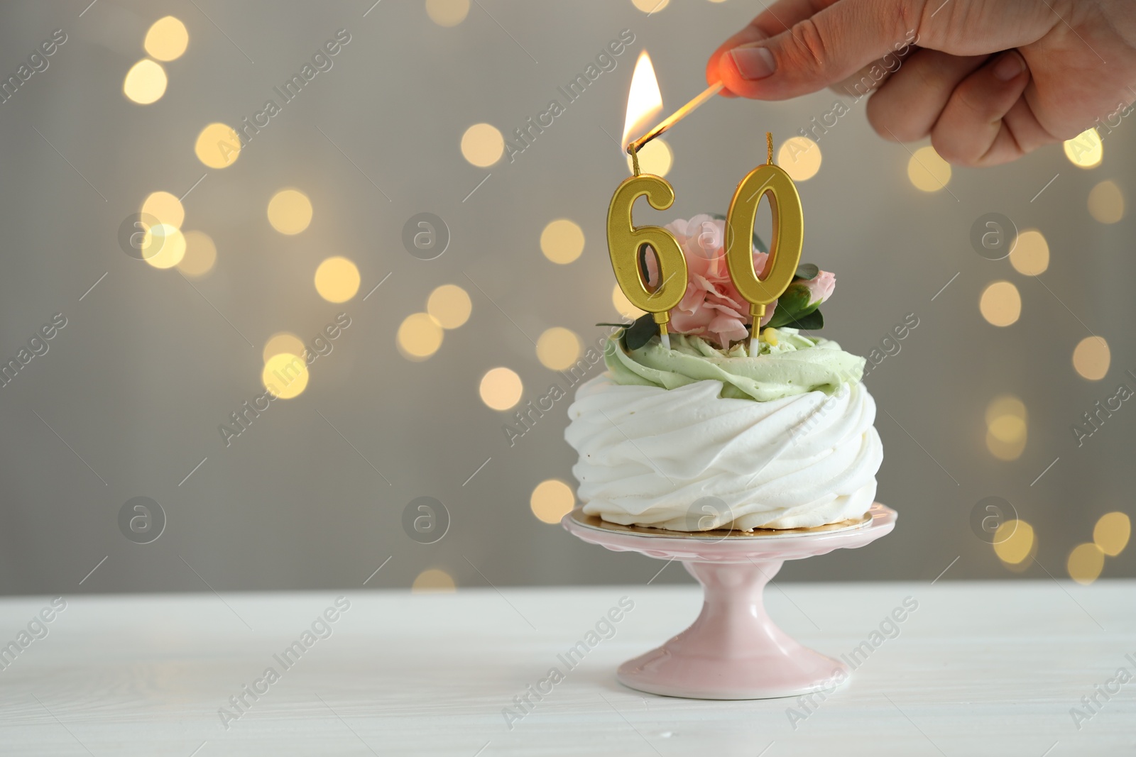 Photo of 60th birthday. Woman lighting number shaped candles on cupcake at white wooden against grey background with blurred lights, closeup. Space for text