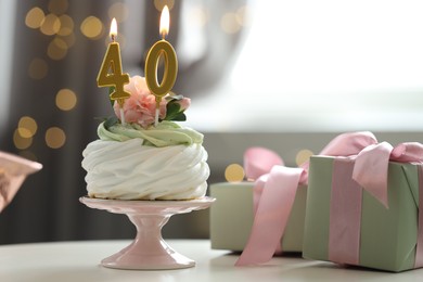 Photo of 40th birthday. Tasty cupcake with burning number shaped candles and gift boxes on table against blurred background with lights, bokeh effect