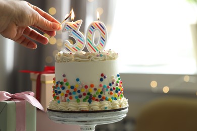 Photo of 30th birthday. Woman lighting number shaped candles on cake against blurred background with lights, closeup. Space for text