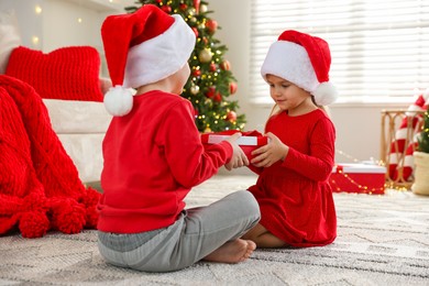 Little girl in Santa hat presenting her brother with Christmas gift at home