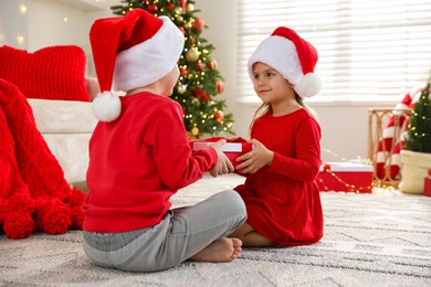 Photo of Little girl in Santa hat presenting her brother with Christmas gift at home