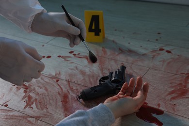 Photo of Forensic expert taking fingerprints with brush from gun and dead woman's body at crime scene indoors, closeup