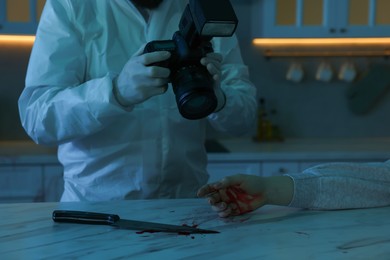 Photo of Forensic expert with camera, knife, blood and dead woman's body at countertop indoors, closeup