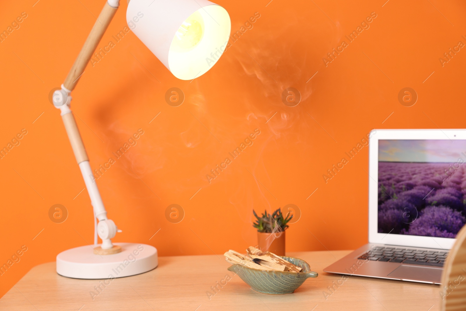 Photo of Palo santo sticks, lamp and laptop on wooden table against orange wall