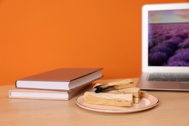 Photo of Palo santo sticks, laptop and books on wooden table against orange wall, space for text