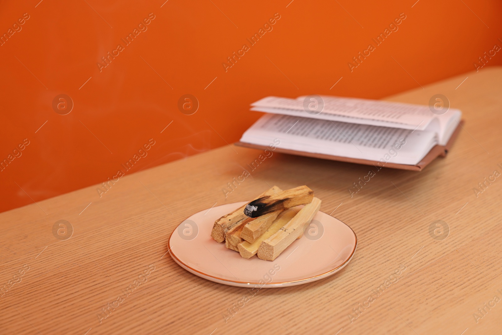 Photo of Palo santo sticks and book on wooden table against orange wall, space for text