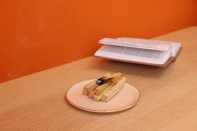 Photo of Palo santo sticks and book on wooden table against orange wall, space for text