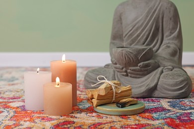 Photo of Palo santo sticks, burning candles and Buddha statue on rug indoors, closeup