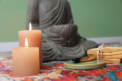 Photo of Buddha statue with palo santo sticks and burning candles on rug indoors, closeup