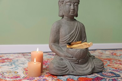 Photo of Buddha statue with palo santo sticks and burning candles on rug indoors