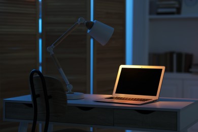 Comfortable workplace with laptop and lamp on table indoors