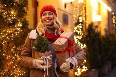 Photo of Happy woman with Christmas gift and thuja tree on city street, space for text