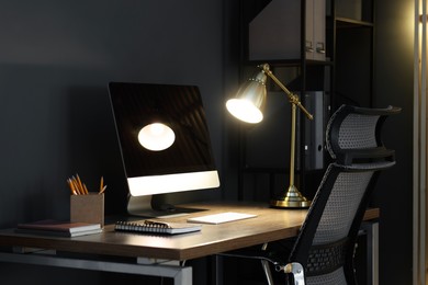 Photo of Comfortable workplace with computer and lamp on wooden desk at home office in evening