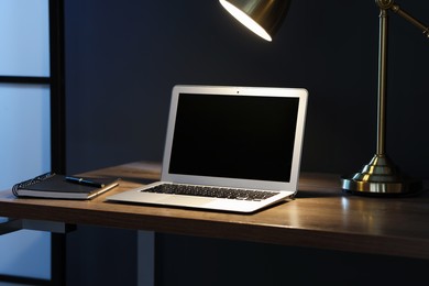 Photo of Comfortable workplace with laptop and lamp on wooden desk at home office in evening