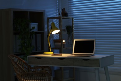 Photo of Comfortable workplace with laptop and lamp on desk at home office in evening