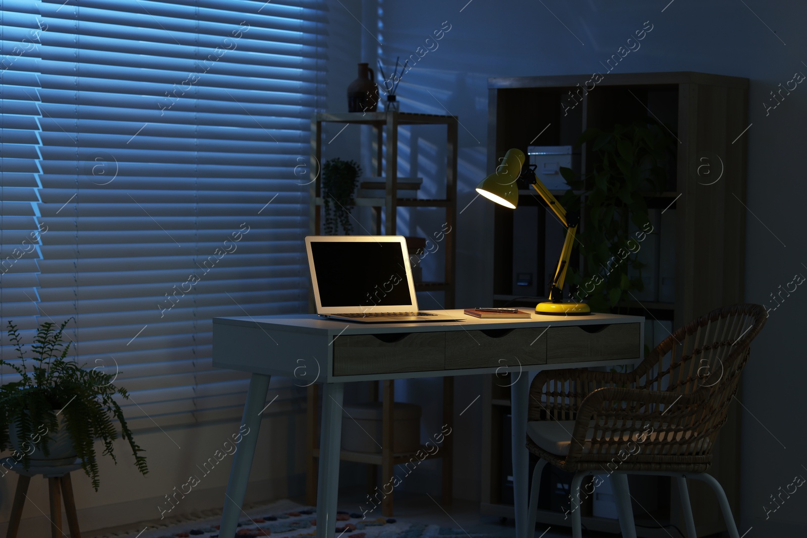 Comfortable workplace with laptop and lamp on desk at home office in evening Photo of Comfortable workplace with laptop and lamp on desk at home office in evening